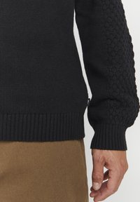 Strikket sweater i sort, med et struktureret mønster på ærmerne, ribbede manchetter og kant, sammen med lysebrune bukser.