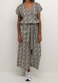 Vestido maxi de tejido beige con patrón abstracto negro, mangas cortas, escote en V, cintura elástica y silueta fluida, combinado con zapatillas blancas.