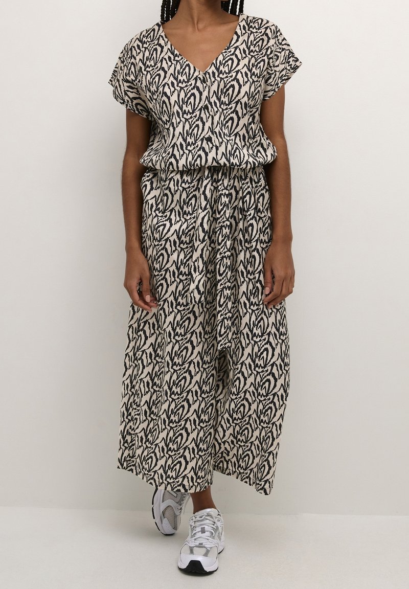 Vestido maxi de tejido beige con patrón abstracto negro, mangas cortas, escote en V, cintura elástica y silueta fluida, combinado con zapatillas blancas.