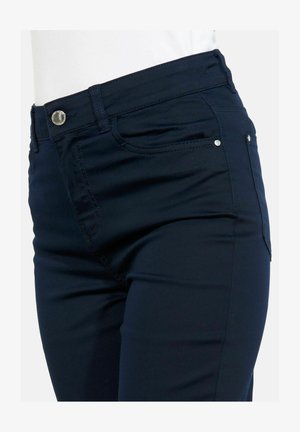 Primo piano di pantaloni aderenti blu scuro con tasche frontali, passanti per cintura e chiusura con bottone argentato su sfondo bianco.