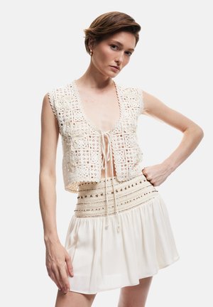 Szydełkowa kamizelka w kolorze off-white z wzorem ażurowym, zapinana na sznurek z przodu, w połączeniu z kremową spódnicą plisowaną z haftowanymi akcentami.