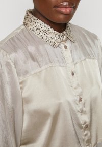 Blusa de color beige claro y suave con un cuello texturizado y adornado con cuentas, y un frente abotonado con un ligero brillo y un patrón de rayas verticales.
