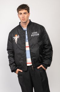 Black bomber jacket with a smooth texture, featuring an embroidered cherub design and “LOVE POTION” text. Worn over a light blue sweater.
