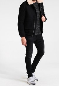 Blouson aviateur noir avec col bordé de fourrure, porté sur un pull gris texturé, associé à un jean noir et des baskets blanches et grises sportives.