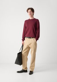 Camicia a maniche lunghe borgogna, pantaloni beige, cintura nera e scarpe in pelle nera. Tiene una borsa a tracolla nera. Sfondo neutro.