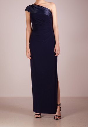 Žena v námořnicky modrých šatech na jedno rameno, dlouhých až po zem s rozparkem, a černých otevřených lodičkách stojící před jednobarevným pozadím.