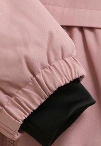 Nærbilde av en rosa jakkeerm med elastisk mansjett over et svart indre stofflag, som viser fiskebensmønster på ytterstoffet.