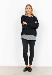 Schwarzes Langarmoberteil, das über einem gestreiften Hemd mit Kragen getragen wird, kombiniert mit schlanken schwarzen Jeans und schwarzen Loafers vor einem neutralen Hintergrund.