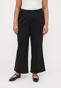 Black wide-leg pants made of soft fabric, featuring a smooth waistband with pleats. Worn with black flat shoes.