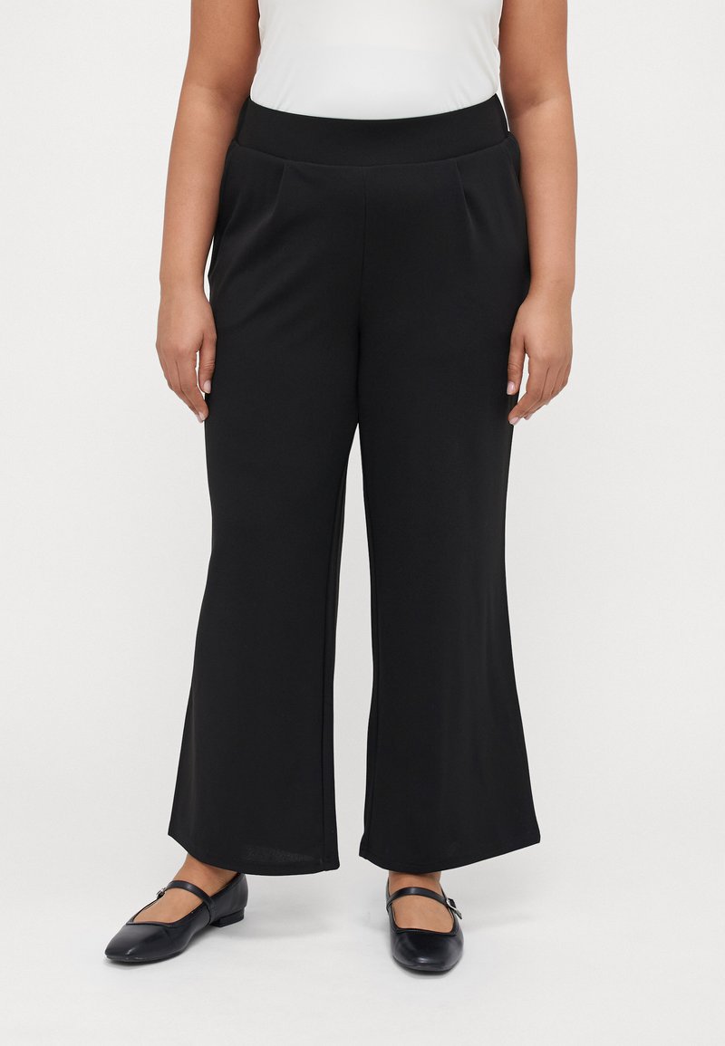 Black wide-leg pants made of soft fabric, featuring a smooth waistband with pleats. Worn with black flat shoes.
