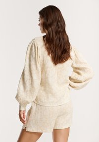 Top beige chiaro con maniche a sbuffo in tessuto testurizzato, caratterizzato da un delicato motivo floreale. Abbinato a pantaloni corti coordinati, anch'essi in beige chiaro.