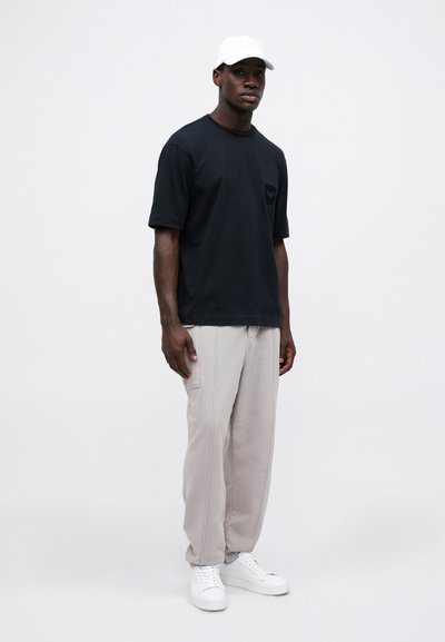 Svart kortermet t-skjorte med brystlomme, kombinert med lysegrå joggebukser og hvite sneakers. Bakgrunnen er enkel hvit.