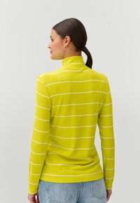 Dolgi zgornji del s turtleneck ovratnikom v svetlo rumeni barvi z belimi vodoravnimi črtami, rebrasto teksturo in prilegajočim se krojem. Ujemanje z modrimi kavbojkami.