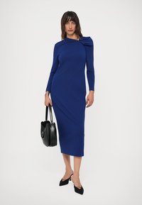Mulher com um vestido midi azul royal de mangas longas e detalhe nos ombros, segurando uma mala preta e usando sapatos de salto pontiagudos pretos.