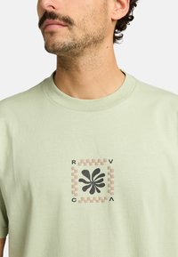 Světlounce zelené bavlněné tričko s centrálním grafickým designem, který zobrazuje černý květinový tvar, obklopený kostkovaným vzorem v růžové a béžové.