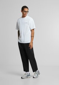 White cotton T-shirt with a blue logo, paired with black loose-fit trousers and gray sports shoes with metallic accents.