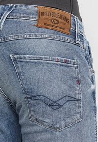 Light blue denim jeans featuring a leather label on the back, two back pockets with a wave pattern and red stitching accents.