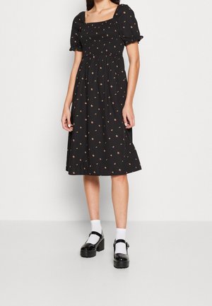 Robe noire avec des manches courtes bouffantes, un corsage smocké et un imprimé floral. Jupe évasée atteignant le mollet. Portée avec des chaussures noires en vernis et des chaussettes blanches.