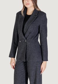 Blazer doppiopetto grigio scuro con tessuto a spina di pesce, rever scollato e dettagli in metallo, abbinato a pantaloni coordinati.