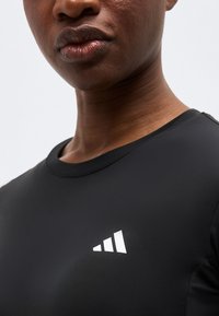 Κοντινό πλάνο ενός ατόμου που φοράει μαύρο πουκάμισο με μικρό λευκό γεωμετρικό λογότυπο στο στήθος.