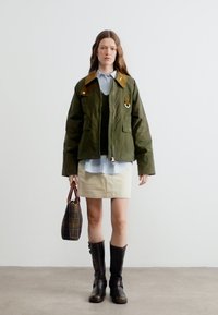 Woman wearing an olive green jacket, striped shirt, beige skirt, black boots, and holding a plaid handbag against a plain white background.