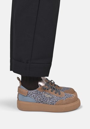 Lage sneakers met luipaardprint, bruine suède accenten, dikke beige zolen en zwarte veters, gedragen met een zwarte broek en zwarte sokken.