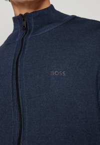 Närbild av en marinblå zip-up-tröja med hög krage och en liten "BOSS"-logotyp på bröstet.