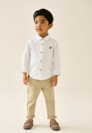 Ung dreng står iført en hvid skjorte med knapper og et lille bjørnepatch, beige optrukne bukser og brune loafers foran en ensfarvet baggrund.