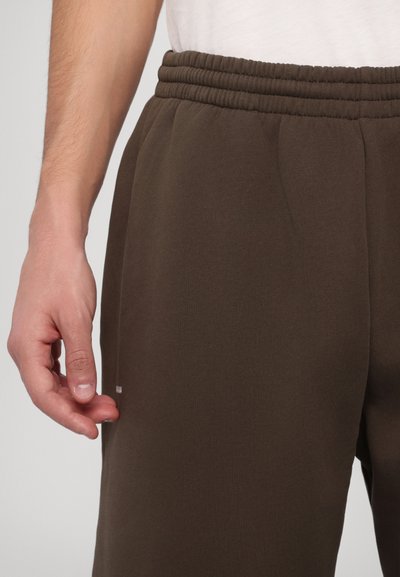 Καφέ φόρμες με ελαστική μέση και πλαϊνές τσέπες. Το ύφασμα είναι λείο και υφασμένο, με ένα διακριτικό λογότυπο της μάρκας κοντά στο στρίφωμα.