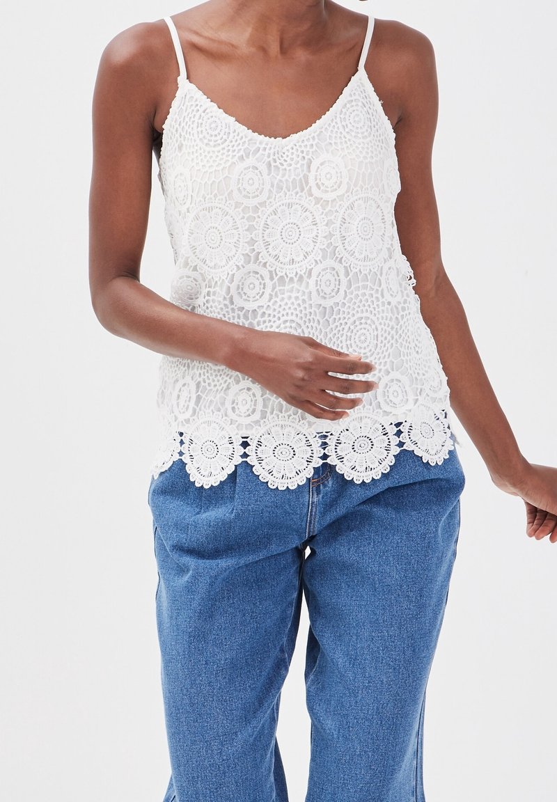 Camisole en dentelle blanche avec un ourlet festonné, présentant des motifs circulaires, des bretelles fines réglables et un tissu léger et texturé.