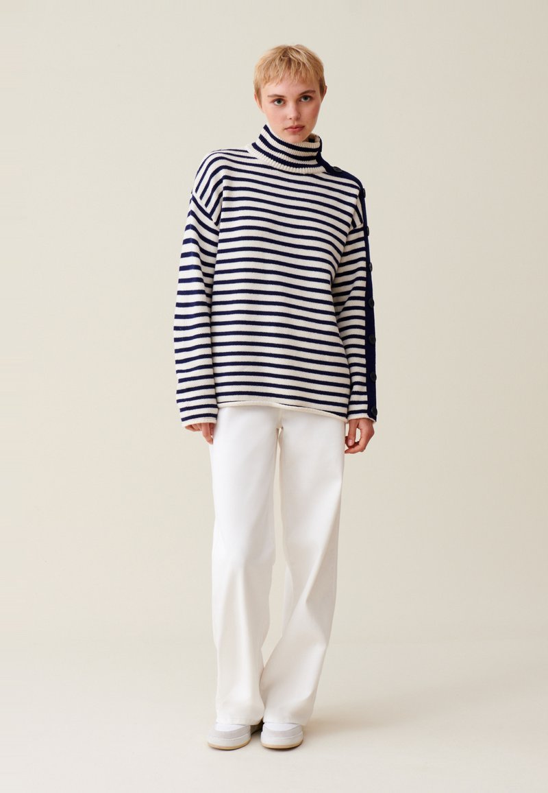 Gestreiftes Rollkragenpullover in Marineblau und Weiß, mit lockerer Passform und Knöpfen an der Seite, kombiniert mit weißen, weiten Hosen.