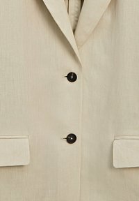Blazer beige avec deux boutons noirs et deux poches à rabat, avec un col à revers cranté.