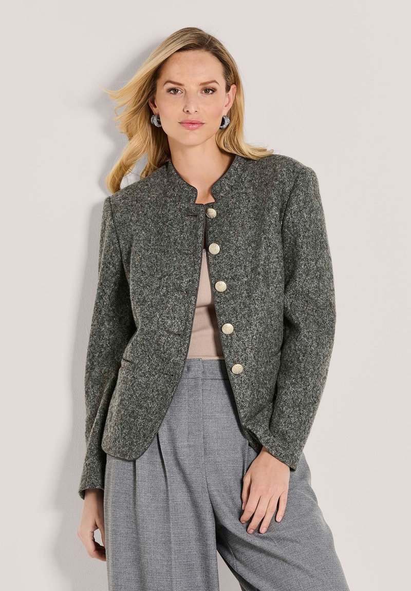 Blonde vrouw in een grijze, getextureerde, knoop-jasje, beige top en grijze wijde broek, poseert met één hand in de zak tegen een effen achtergrond.