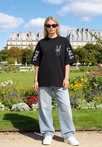 Femme portant un T-shirt oversize noir avec "Take Care" sur les manches et un graphisme main de la paix, jean bleu clair et baskets blanches dans un parc ensoleillé.