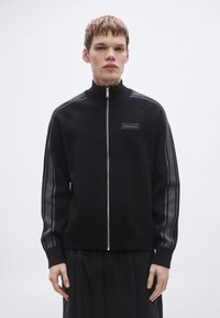 Zwarte jas met ritssluiting, gemaakt van zachte stof, met een hoge kraag, grijze strepen aan de zijkanten en een Calvin Klein-logo op de borst.