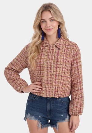 Blonde Frau trägt eine strukturierte rosa-beige Langarmjacke, blaue Quastenohrringe und ausgefranste Jeansshorts, lächelt mit einer Hand in der Taille.