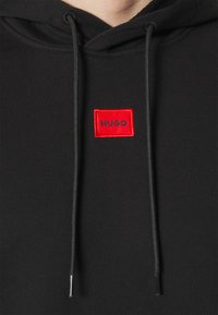 Schwarzer Hoodie mit einem roten rechteckigen Logopatch mit der Aufschrift „HUGO“ in schwarzer Schrift, verstellbaren Kordeln und weicher Stoffstruktur.