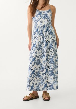 Kobieta ubrana w białą, bezrękawnikową sukienkę maxi z niebieskimi kwiatowymi wzorami i brązowe sandały z klamrą, stojąca na jednolitym tle.