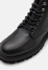 Botte de combat en cuir noir avec bout rond, œillets en métal, lacets noirs et semelle à crampons robuste sur fond blanc.