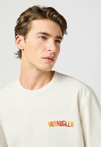 Camiseta blanca de algodón con un logo "WRANGLER" multicolor en naranja y amarillo. Cuello redondo simple y mangas cortas.
