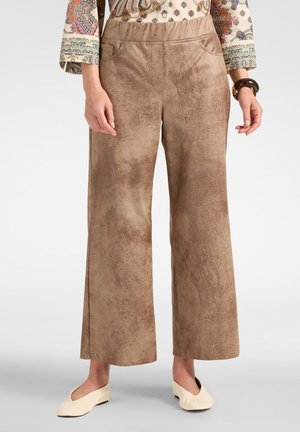Pantalon large en daim marron porté avec un haut à manches longues à motifs, des ballerines crème et un bracelet foncé au poignet droit.