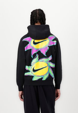 Personne aux cheveux tressés portant un sweat à capuche noir avec des motifs abstraits colorés de soleil et des logos Nike swoosh dans le dos.