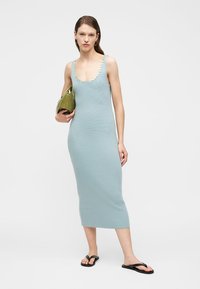 Femme portant une robe midi bleu clair côtelée sans manches avec des embellissements ronds argentés, tenant une pochette vert olive, et des tongs noires.