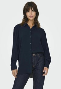 Navy blue long-sleeve button-up shirt with a textured fabric and a classic collar, paired with dark denim jeans featuring visible stitching.