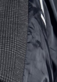 Schwarze Jacke mit strukturierter Hahnentritt-Musterung auf einer Seite und glatter Oberfläche auf der anderen. Kontrastierende Nähte sind sichtbar.