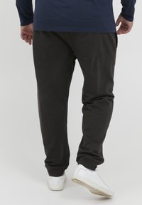 Sorte joggebukser laget av mykt materiale med en innsnevret ben design, inkludert baklommer og ribbede mansjetter, iført med hvite joggesko.
