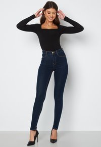 Černý dlouhý rukáv bodysuit s čtvercovým výstřihem spárovaný s tmavě modrými slim-fit džíny s vysokým pasem, které mají zlaté knoflíky a viditelné švy.