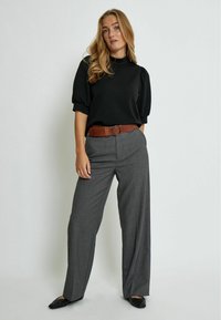 Haut noir à manches bouffantes associé à un pantalon large gris. Ceinture marron avec une boucle ronde. Des chaussures plates noires complètent la tenue, le tout sur un fond neutre.