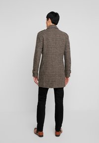 Abrigo de cuadros marrón con un diseño estructurado, mangas largas, cuello y bolsillos laterales. Lleva puesto sobre pantalones negros y zapatos marrones.