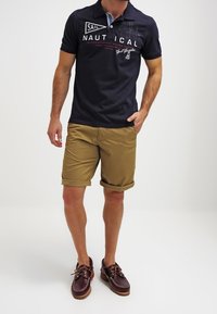 Polo-shirt marine avec col et logo, associé à un short kaki et des chaussures bateau marron. Le short est retroussé à l'ourlet.